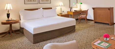 A hotel room with a bed, two bedside tables with lamps, a desk with a chair, a small table with a book and a vase of flowers, and a cabinet.