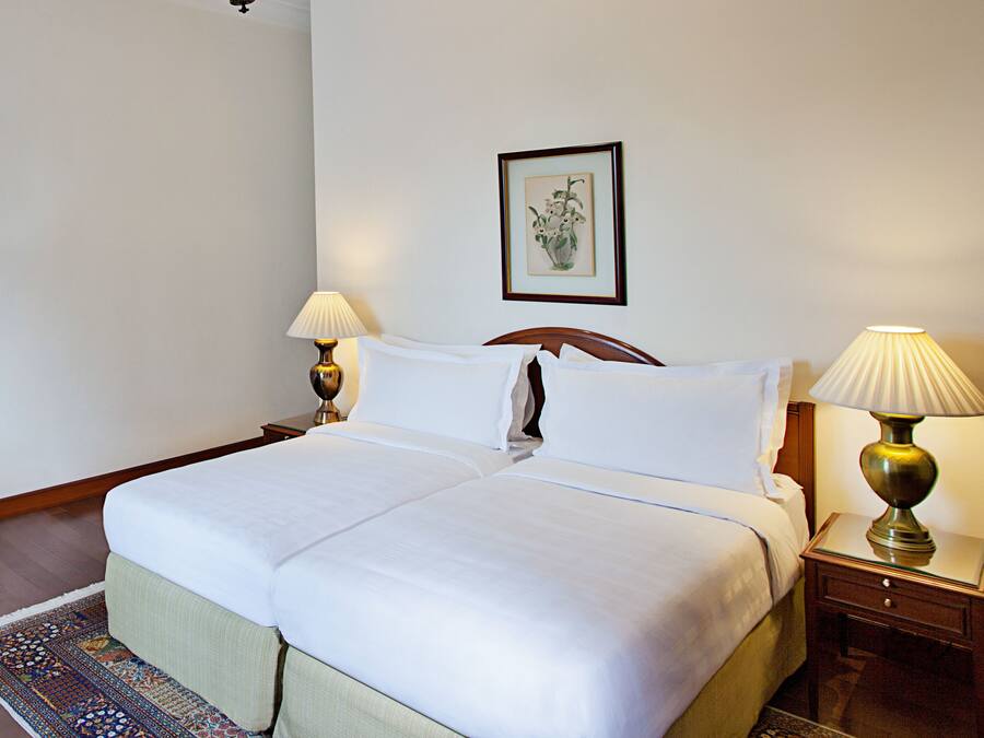 A neatly made bed with white linens, two bedside lamps, a framed floral artwork on the wall, and a patterned rug on the floor.