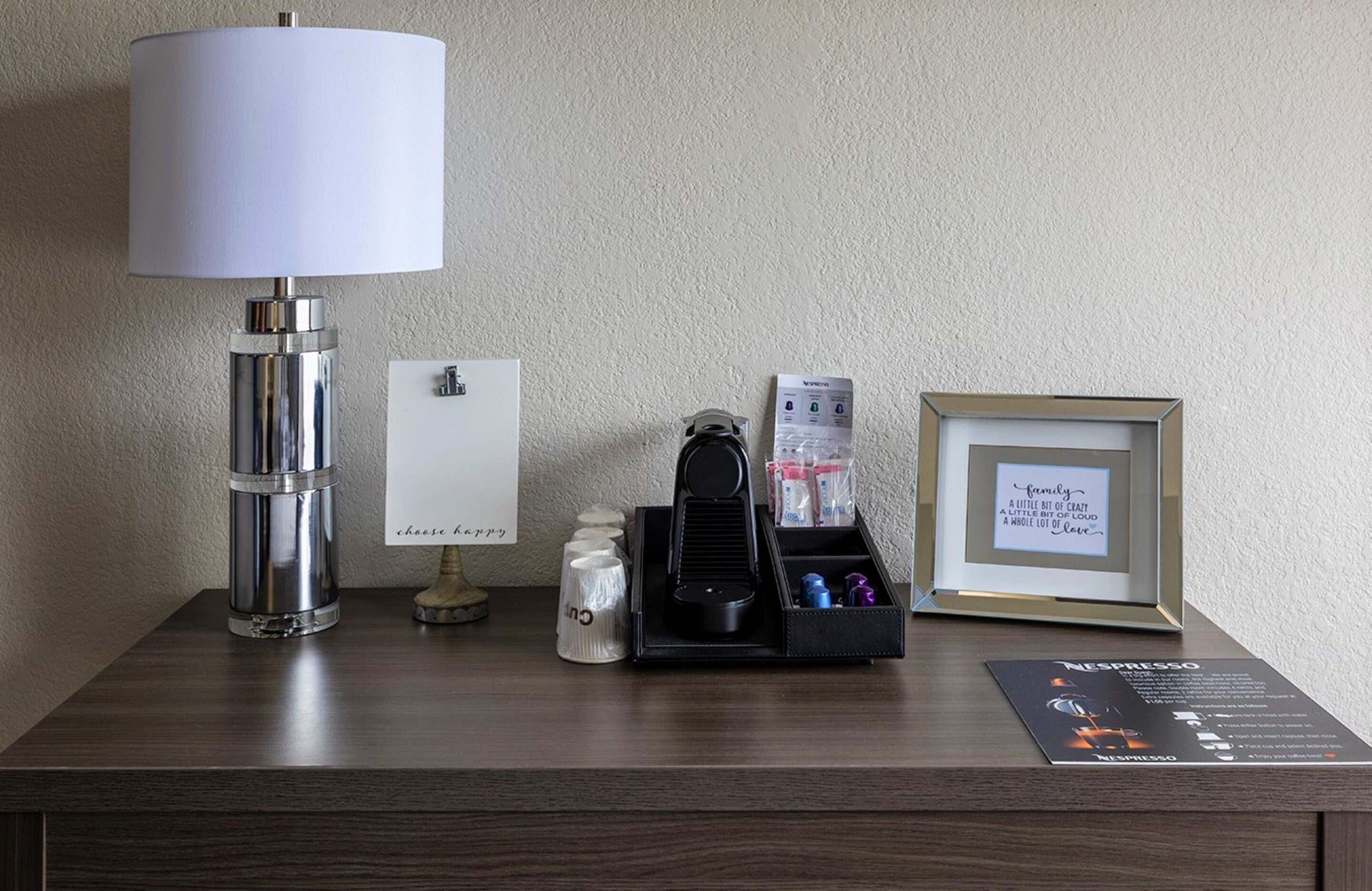 Habitación de hotel con una lámpara, una cita enmarcada, una cafetera y una máquina de Nespresso sobre la mesa.