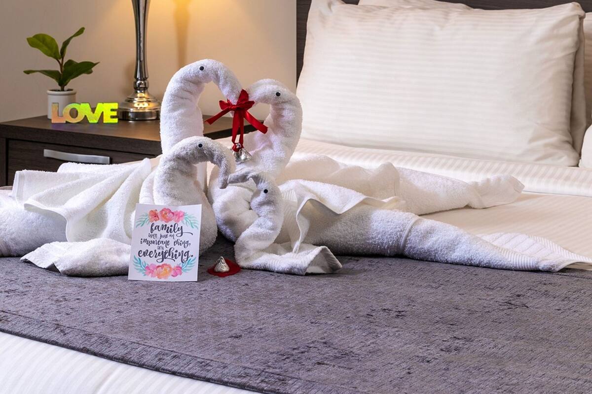 A hotel room with a neatly made bed, a bedside lamp, a decorative towel arrangement, and a card with a family message.