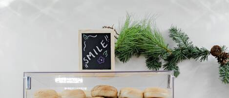A bakery display case with two shelves of baked goods, a chalkboard sign above, and a small potted plant to the side.