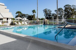 Une piscine entourée d’une clôture blanche et de palmiers, avec des chaises longues et des parasols.