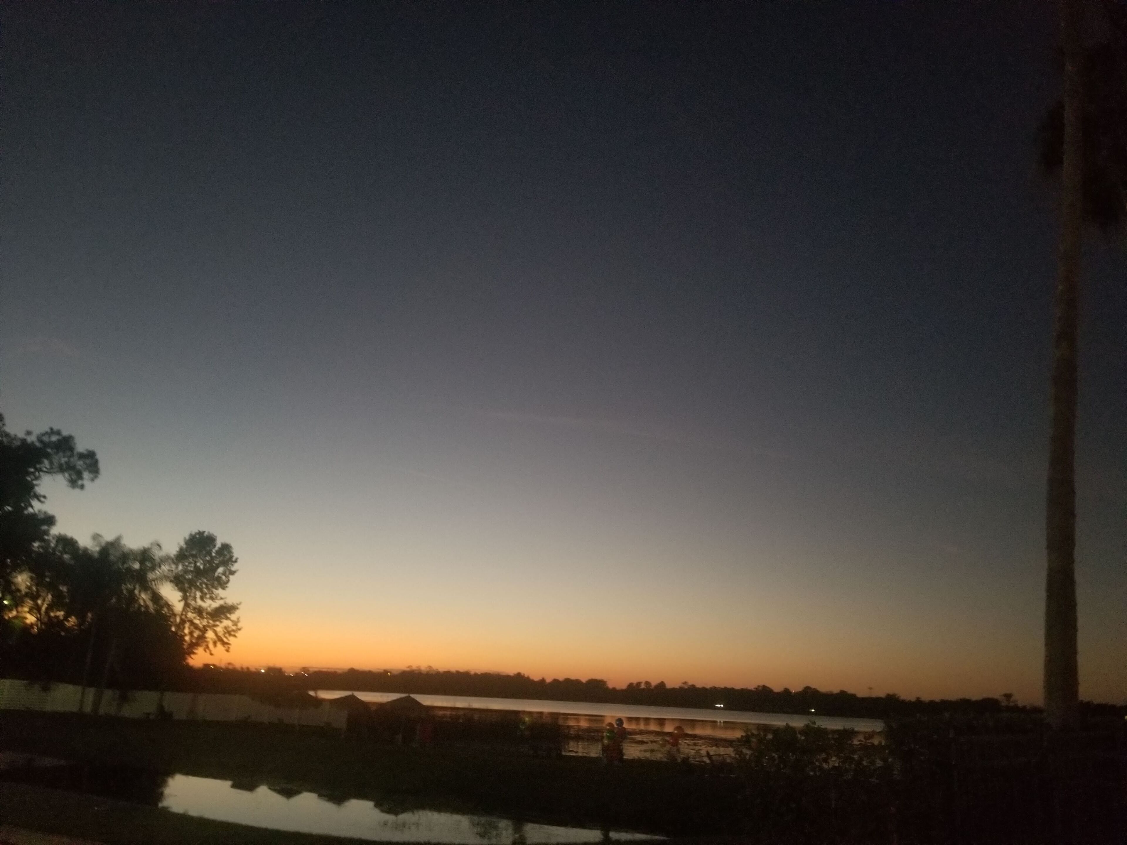 A sunset over a calm body of water with a distant shoreline and a tall structure on the right.