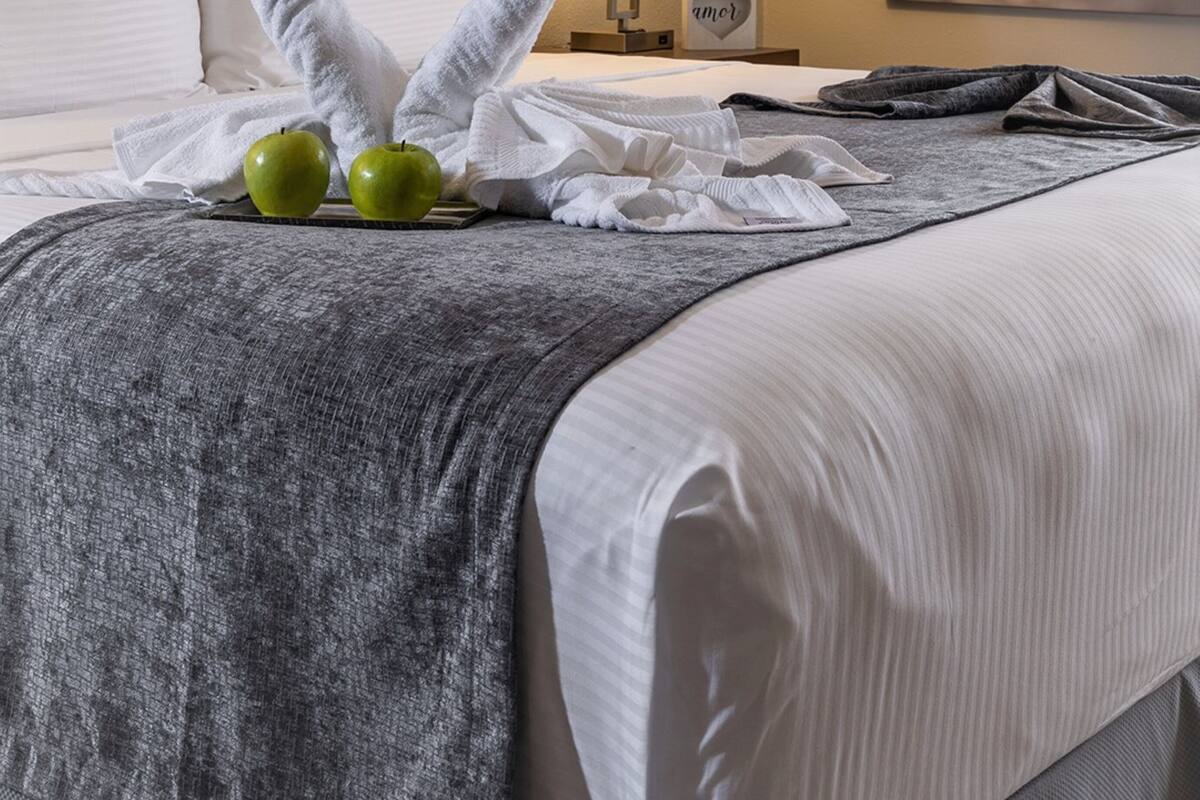 A neatly made bed with a heart-shaped towel and apples, a bedside lamp, and a framed picture on the wall.