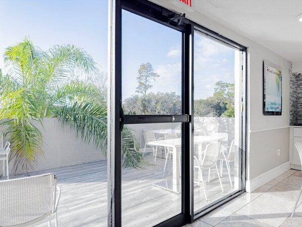 A modern room with a large sliding glass door leading to an outdoor patio with white chairs and tables, surrounded by lush greenery. - Palazzo Lakeside Hotel (Kissimmee)
