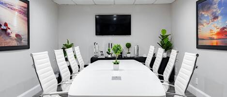 Een vergaderzaal met een lange tafel, witte stoelen, een flatscreen televisie en ingelijste foto's aan de wanden.