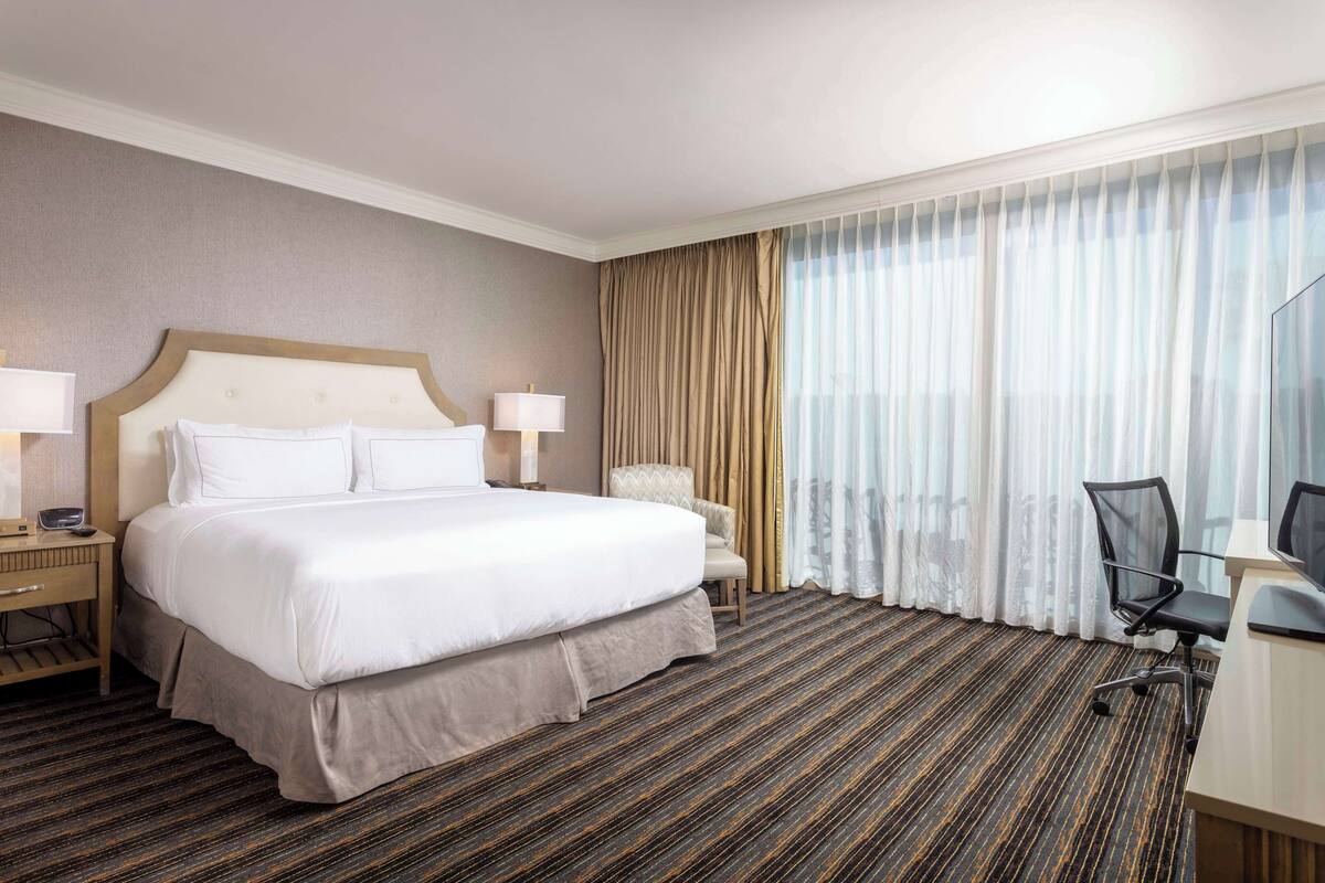 A hotel room with a large bed, a desk with a chair, a lamp, and a view of the city through sheer curtains.