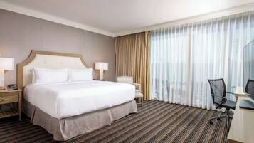 Habitación de hotel con una cama grande, un escritorio con silla, una lámpara y vistas de la ciudad a través de cortinas translúcidas.