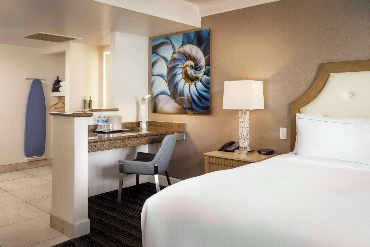 A hotel room with a bed, a desk with a chair, a nightstand, a wall-mounted lamp, and a large abstract painting.