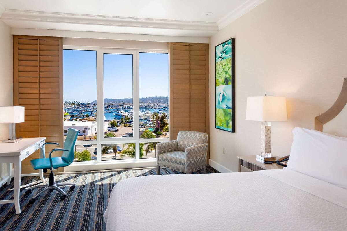 A hotel room with a large bed, a desk, a chair, and a view of a cityscape through large windows.
