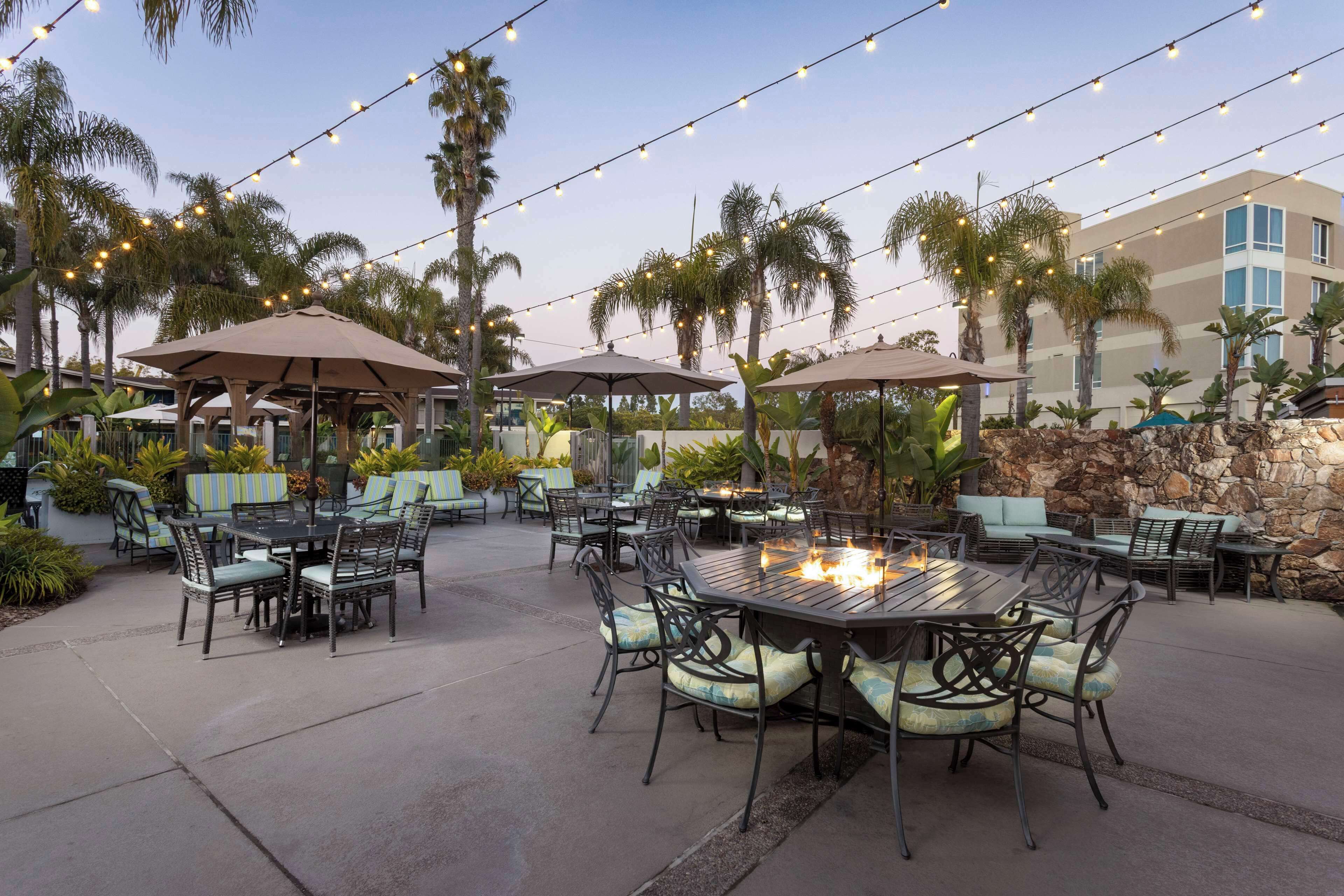 An outdoor patio area with tables and chairs, string lights, and palm trees.