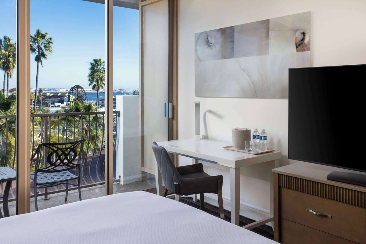 A hotel room with a balcony, a desk, a chair, a bed, a TV, and a minibar.