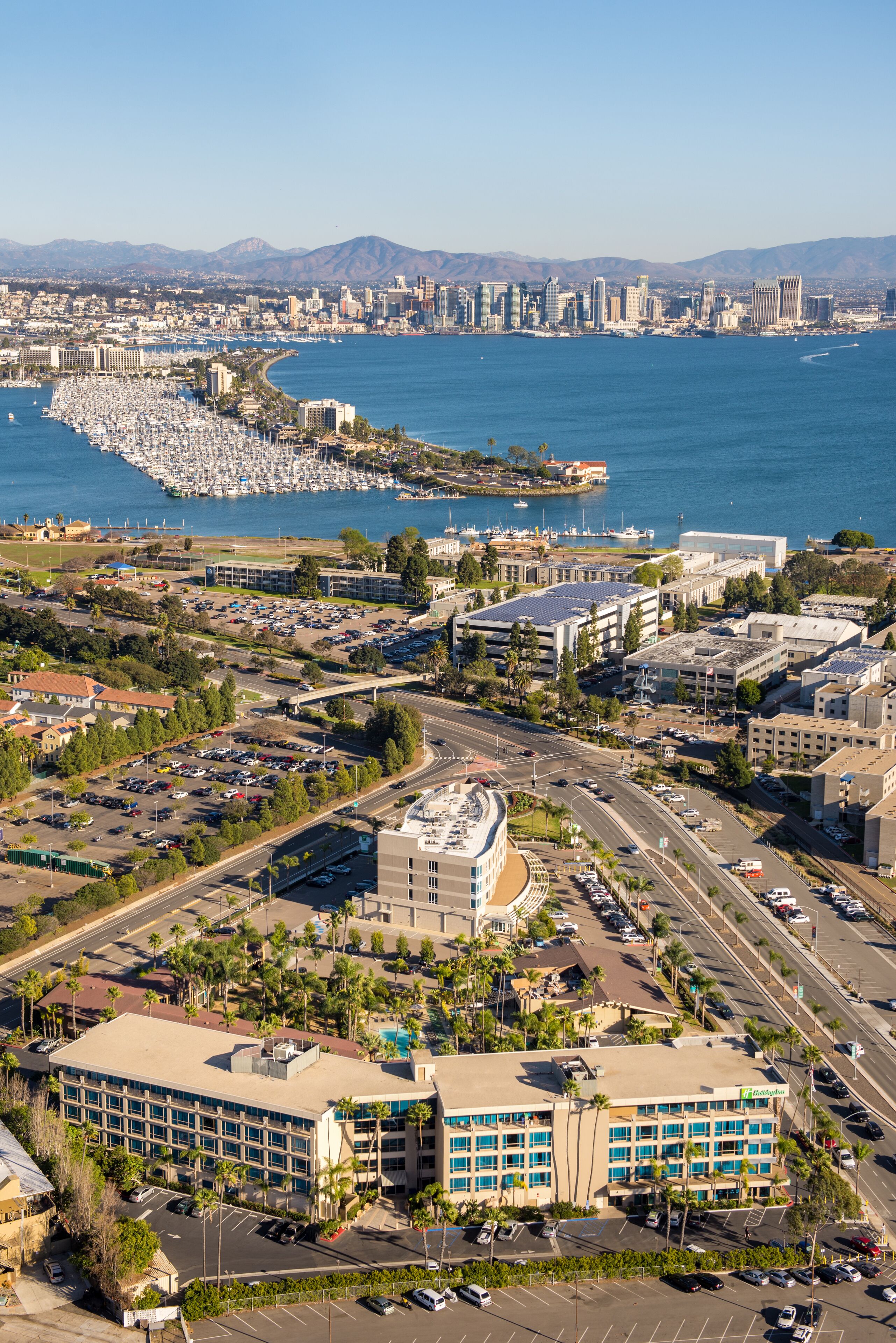 A high-rise hotel with a parking lot, surrounded by palm trees and a marina with boats.