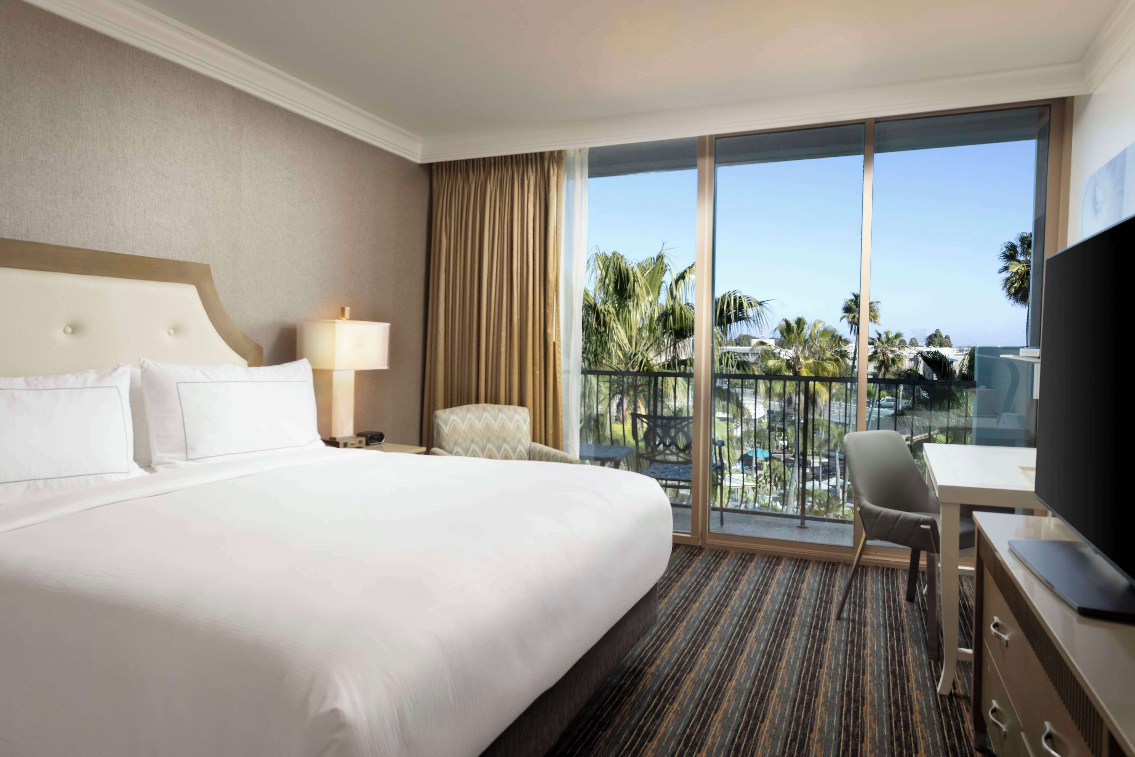 A hotel room with a large bed, a desk, a chair, a TV, and a balcony with a view of palm trees and a pool.