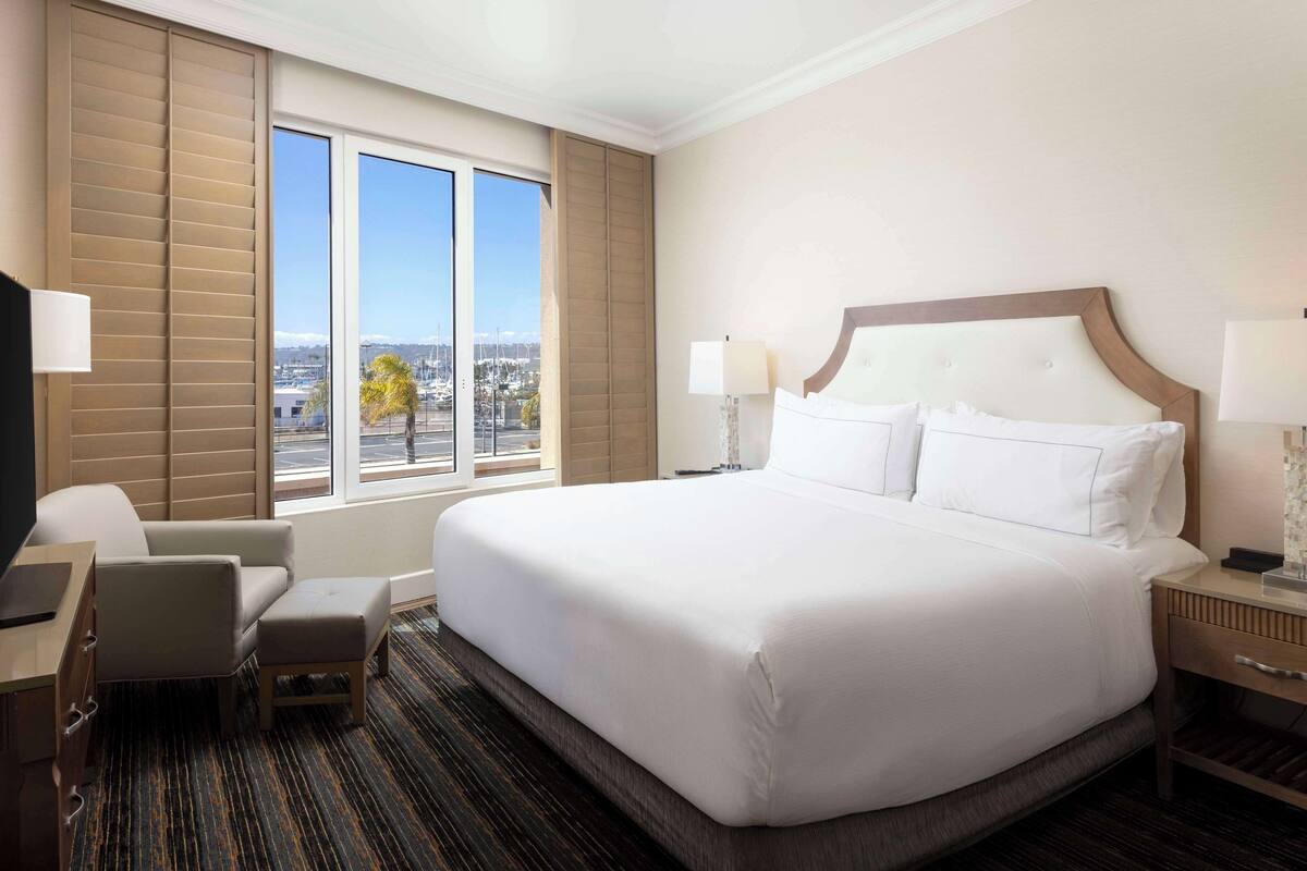 A hotel room with a large bed, a desk, a chair, and a view of the city through large windows.