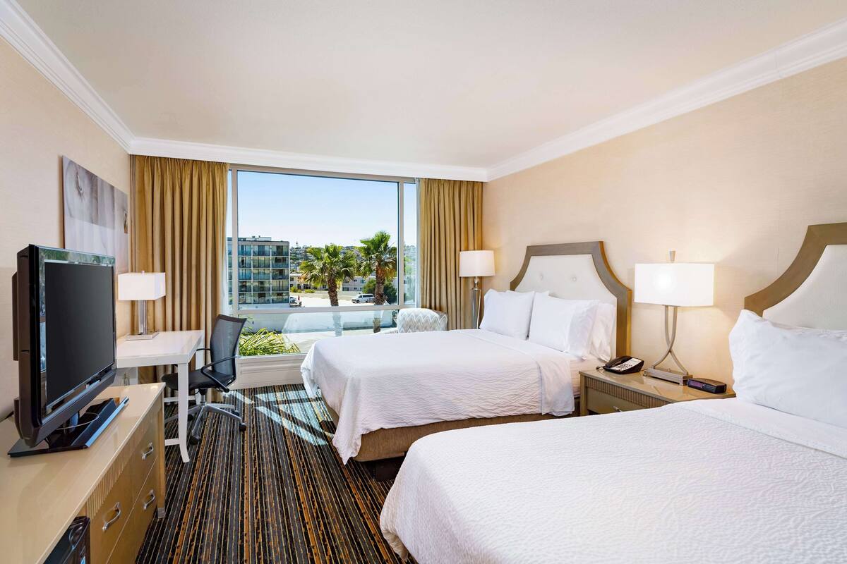 A hotel room with two beds, a desk with a computer, a TV, and a window with a view of palm trees and buildings.