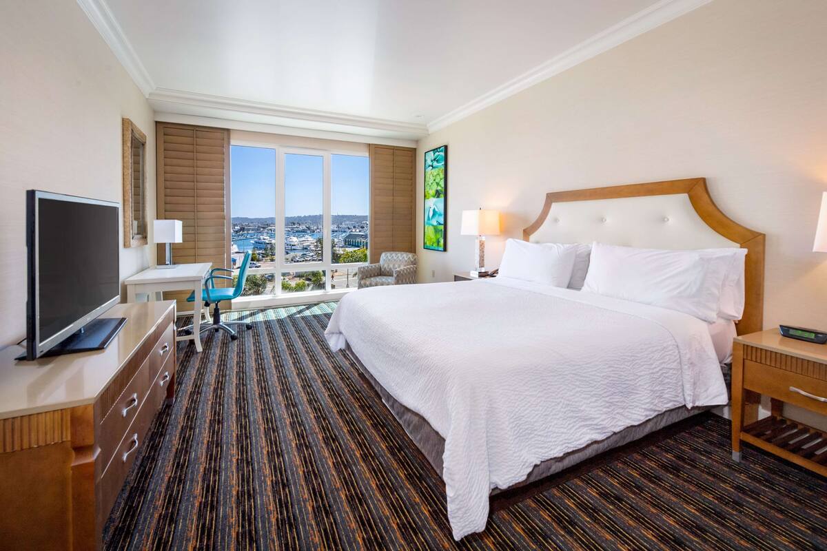 A hotel room with a large bed, a TV, a desk with a chair, and a view of the city.