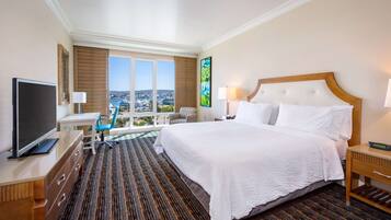 A hotel room with a large bed, a TV, a desk with a chair, and a view of the city.