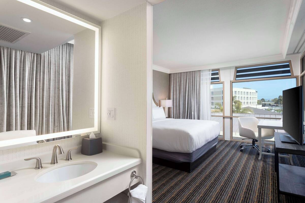 A hotel room with a large bed, a desk, a chair, a TV, and a bathroom with a sink and mirror.