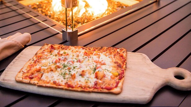 Une pizza carrée servie sur une planche en bois, avec un foyer en arrière-plan.