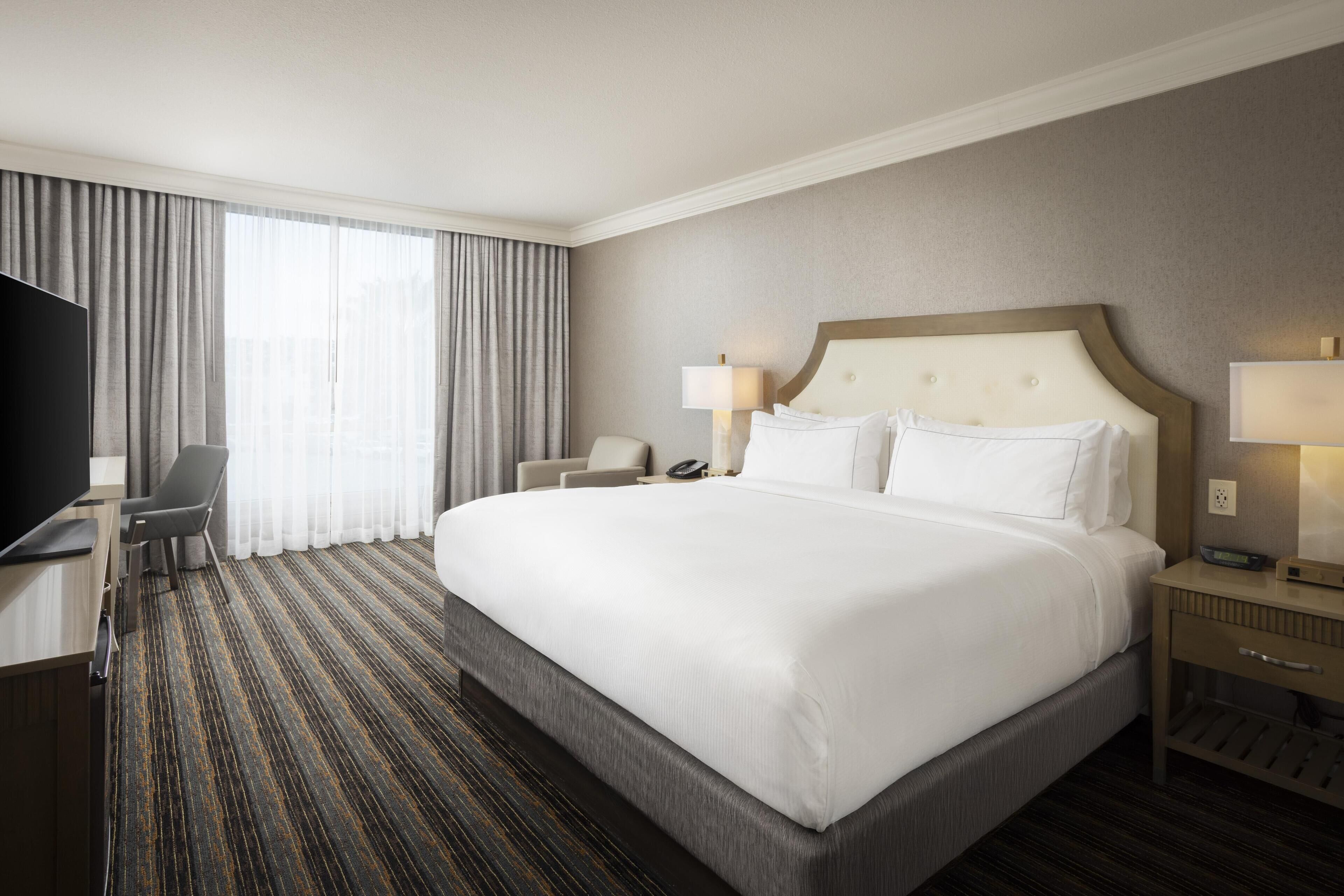 A hotel room with a large bed, a desk with a flat-screen TV, a chair, a bedside table with a lamp, and a window with curtains.