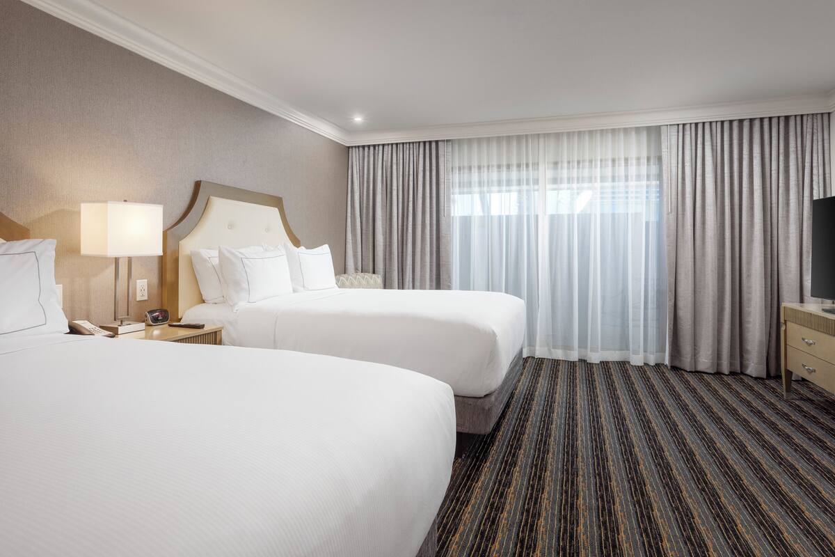 A hotel room with two beds, a TV, a lamp, and a large window with curtains.