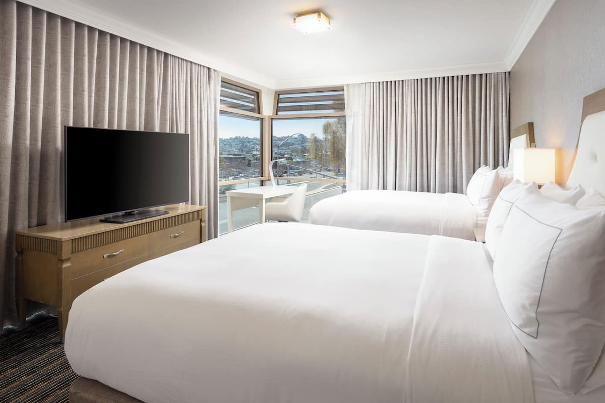 A hotel room with two beds, a flat-screen TV, a desk, and large windows with curtains.