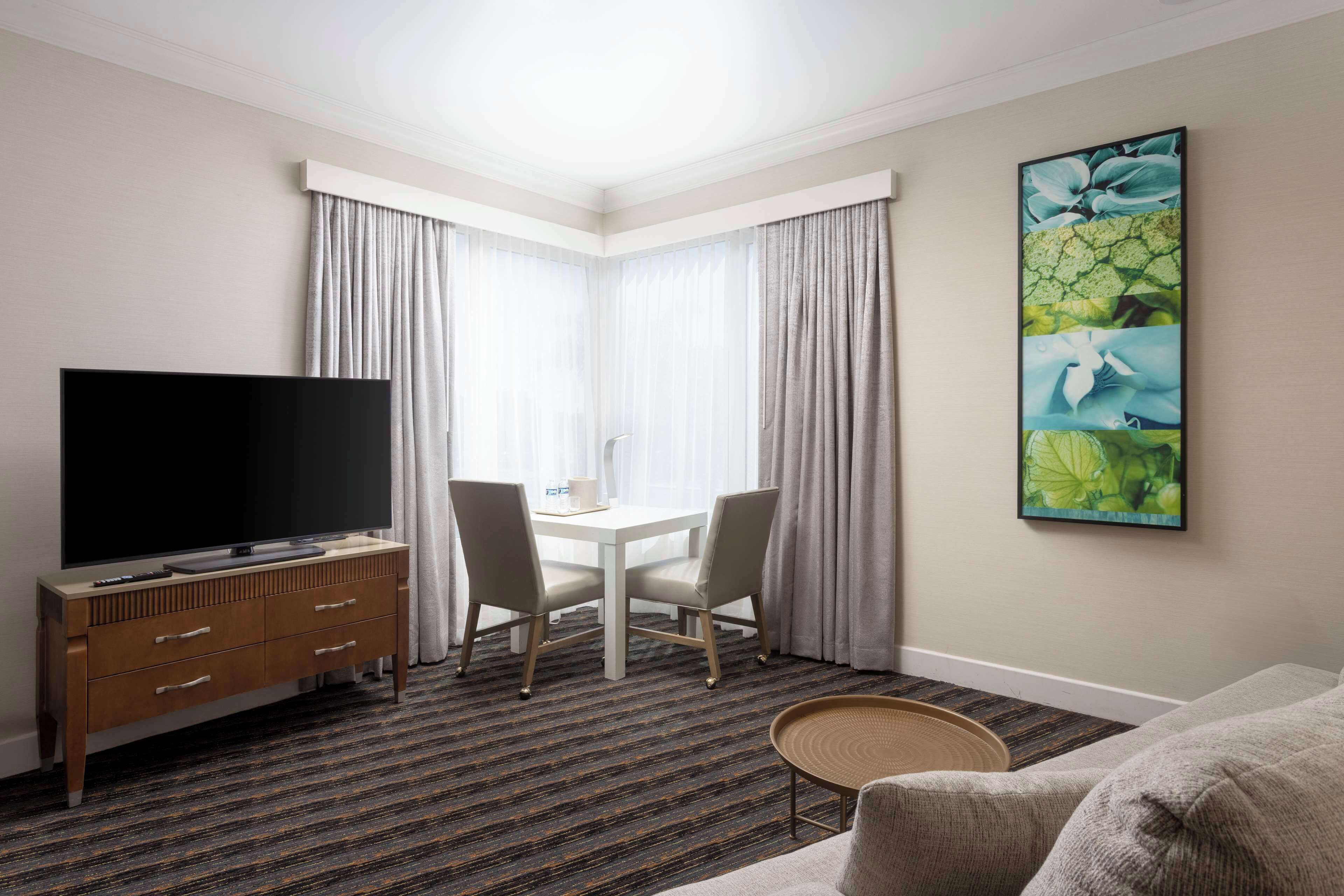 A hotel room with a flat-screen TV, a dining table with chairs, a sofa, and a large floral artwork on the wall.