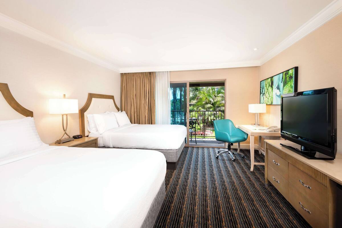 A hotel room with two beds, a desk with a TV, and a view of a garden.