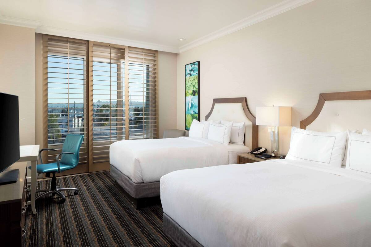 A hotel room with two beds, a desk, a chair, and a large window with blinds.