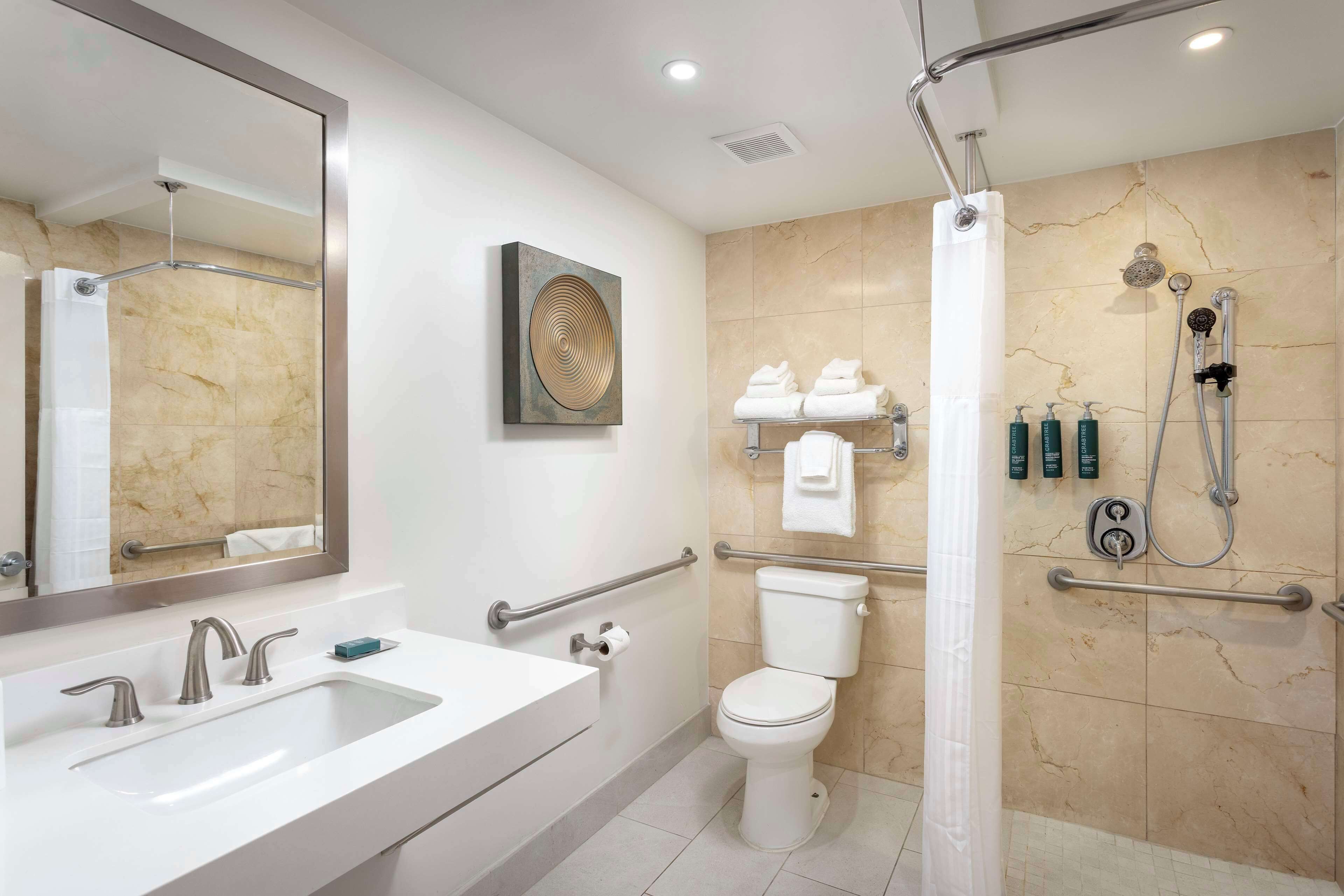 A modern bathroom with a large mirror, dual sinks, a toilet, a shower with a handheld showerhead, and a towel rack with folded towels.
