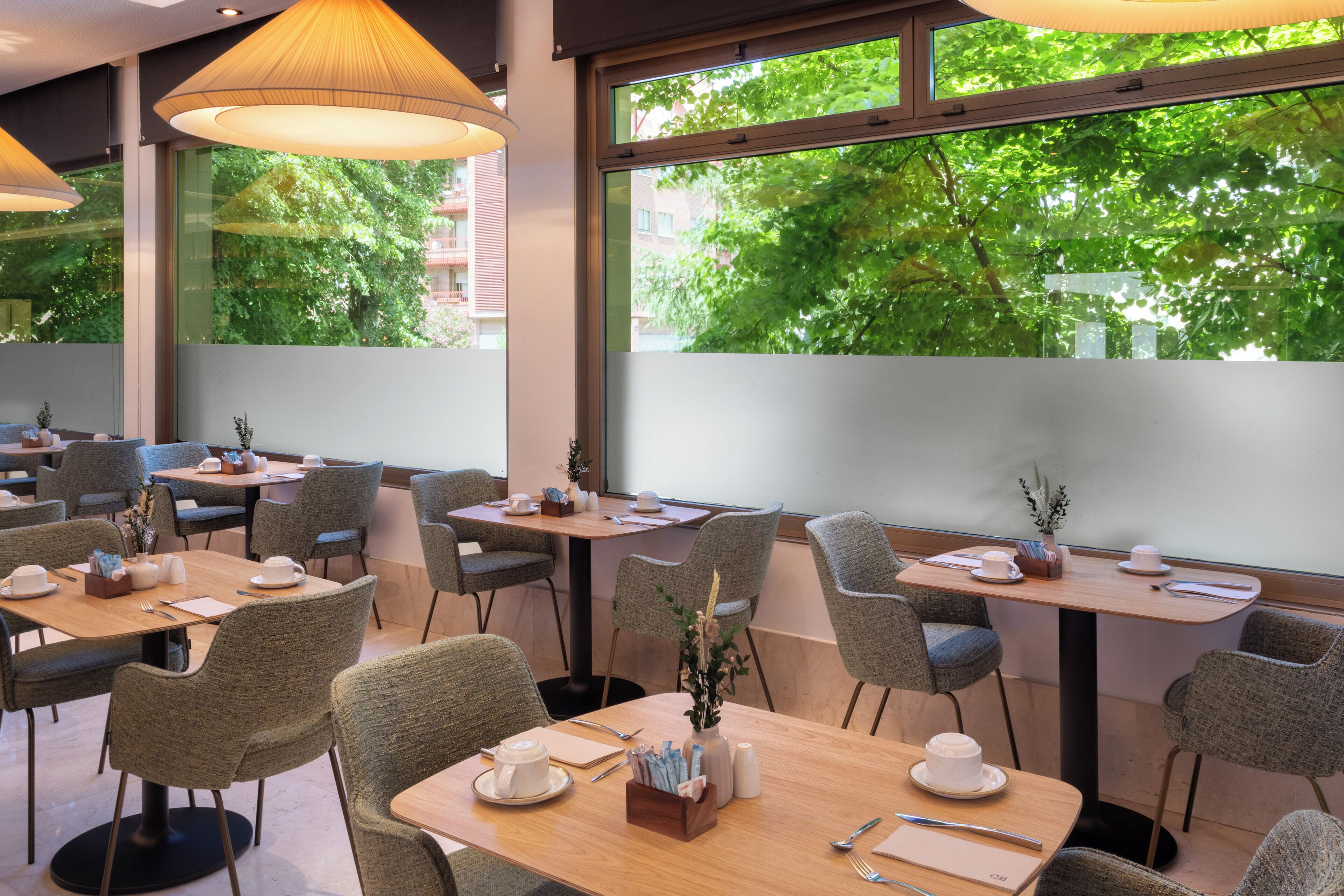 Un espace de restauration moderne avec des tables et des chaises, de grandes fenêtres donnant sur la verdure et des luminaires suspendus.