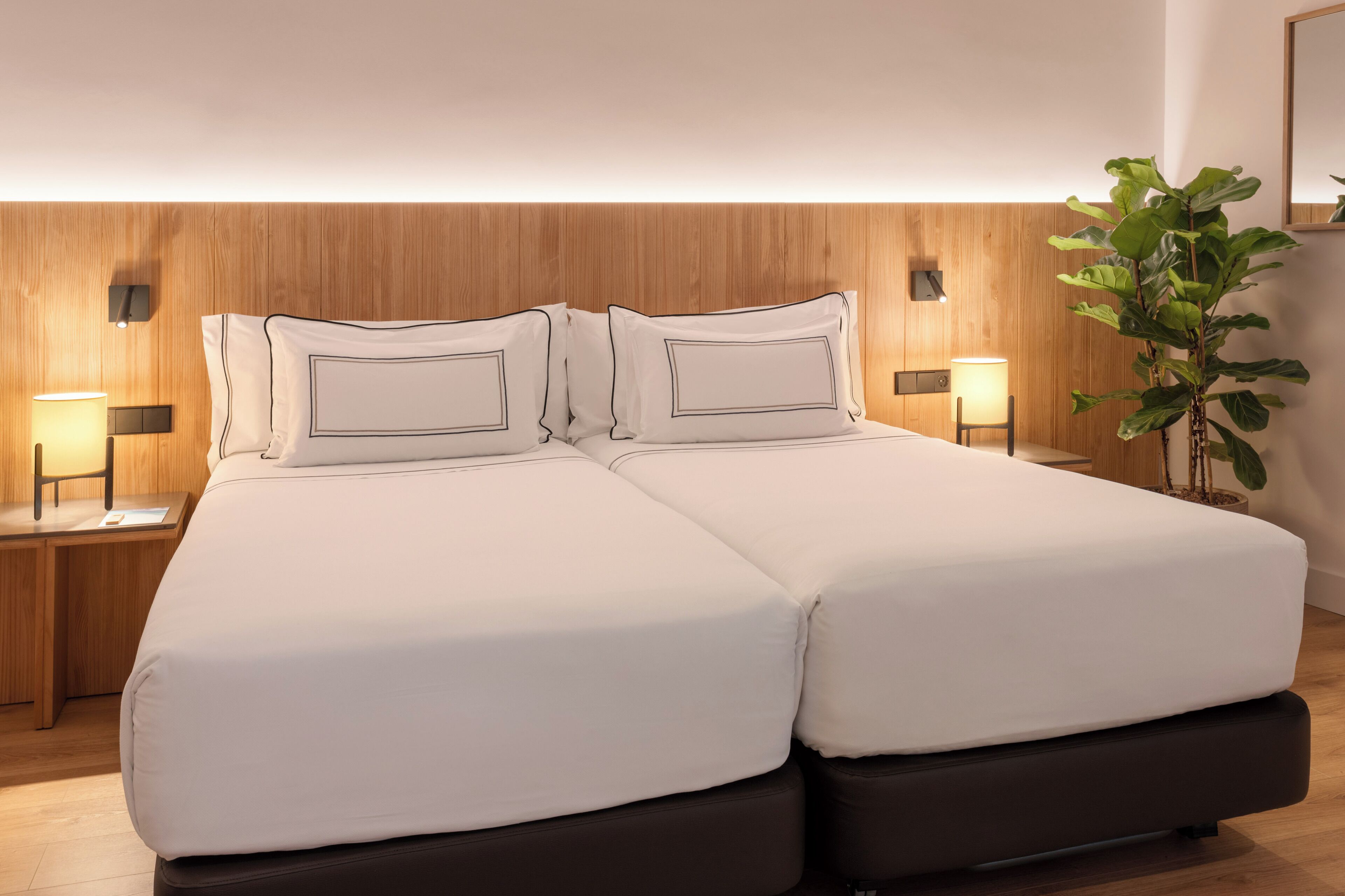 Una cama doble con sábanas blancas y cabecera de madera. Hay dos mesitas de noche con lámparas, una planta en maceta y un espejo en la pared.