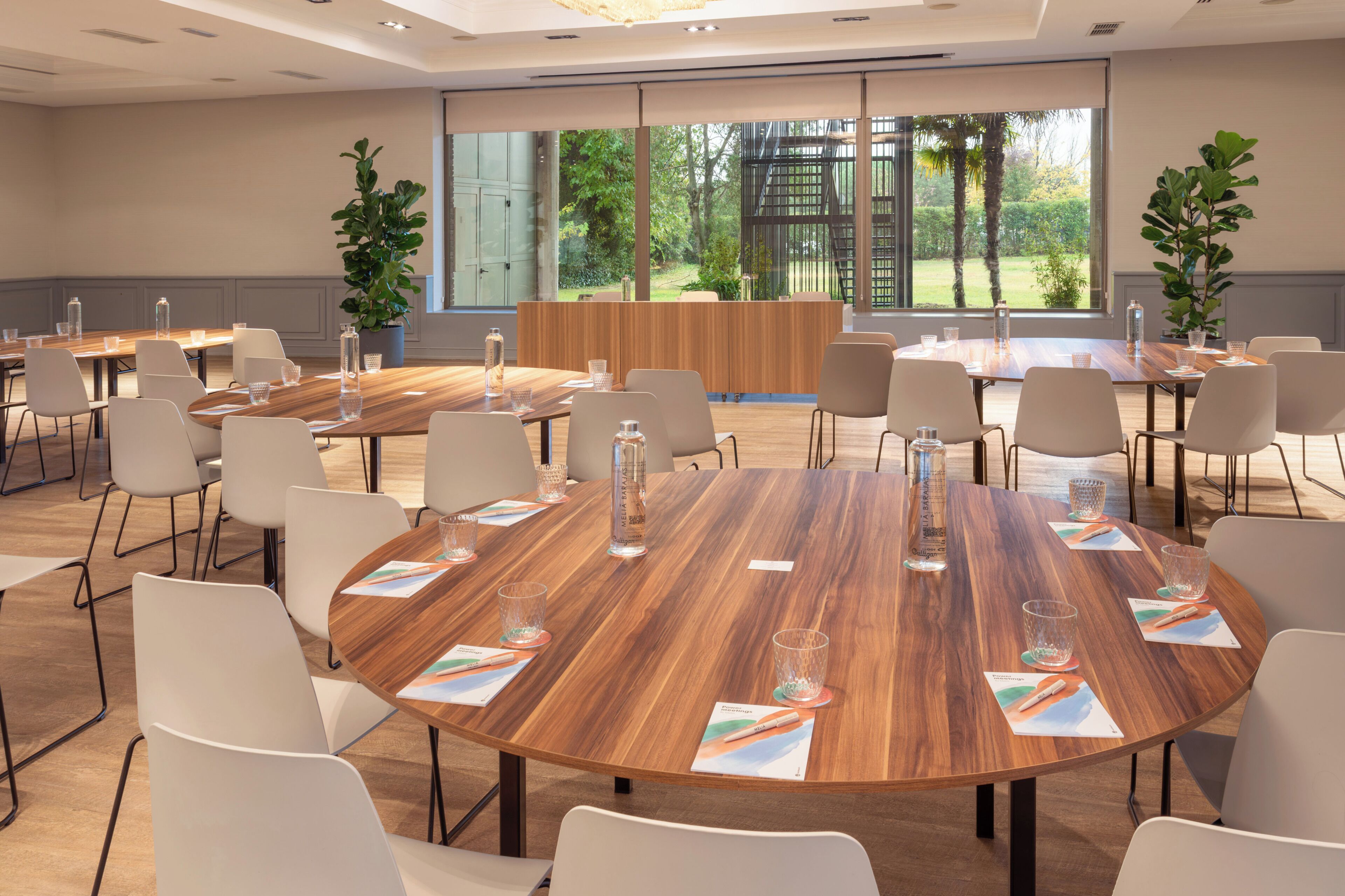 Un salón de conferencias con varias mesas, sillas blancas y ventanas amplias con vistas a zonas verdes.