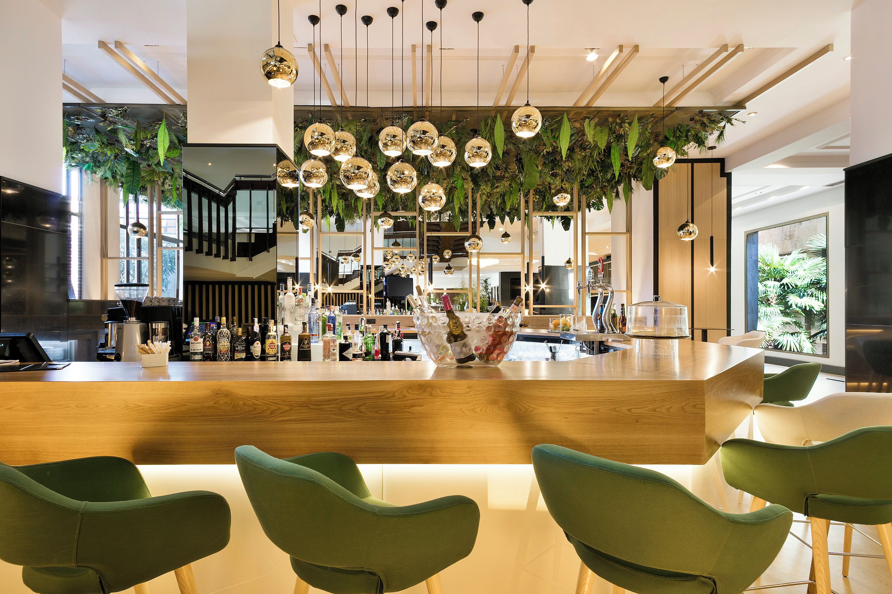 Un área de bar moderna con mostrador de madera, sillas verdes y lámparas esféricas colgantes.