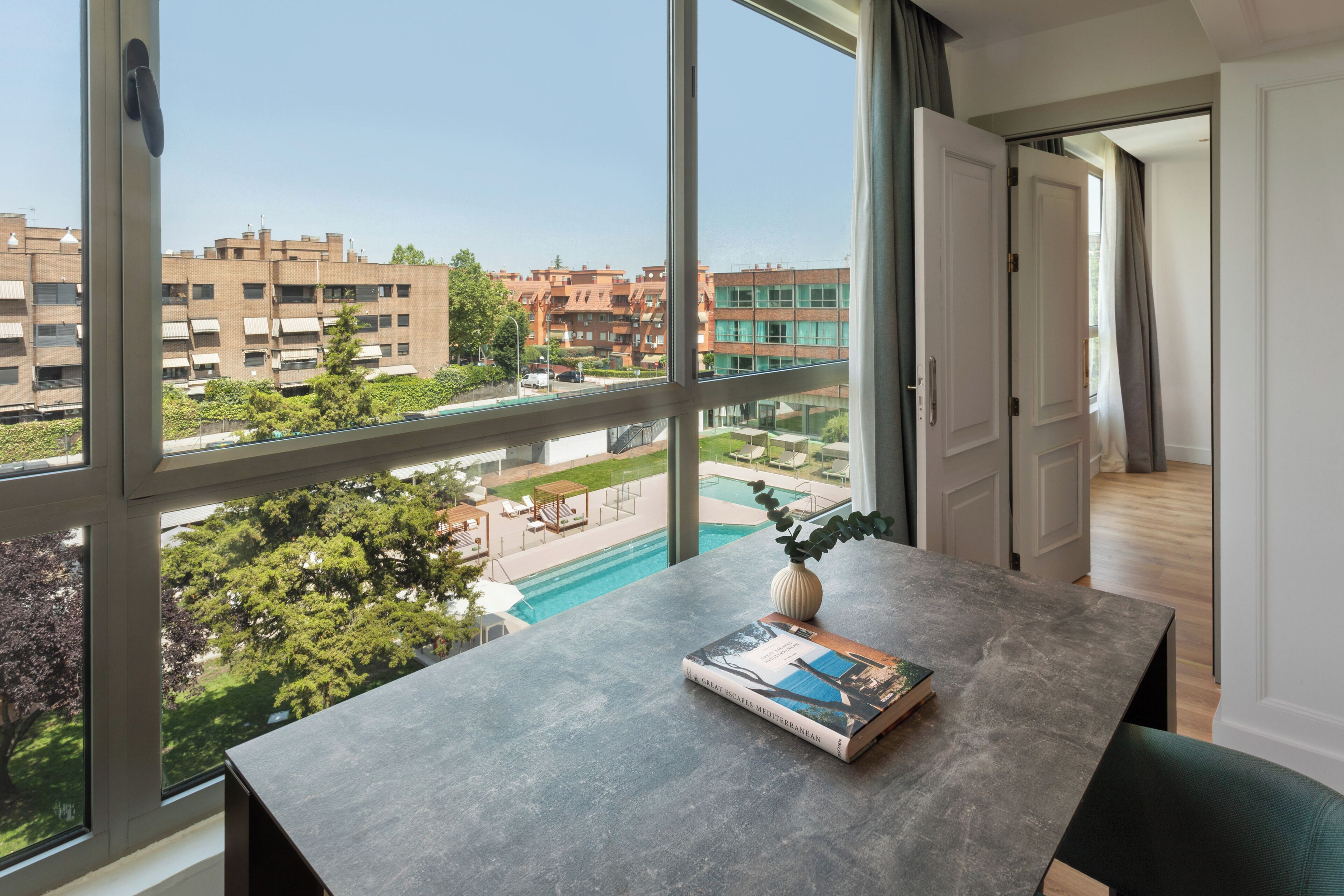 A modern apartment with a large window overlooking a swimming pool and greenery.