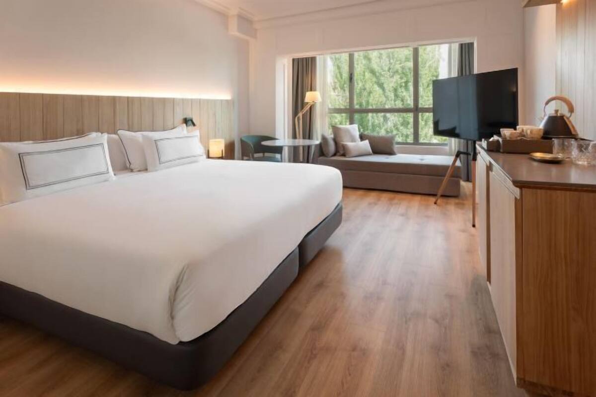 A hotel room with a large bed, a sofa, a small table, a television, and a window with a view of greenery.