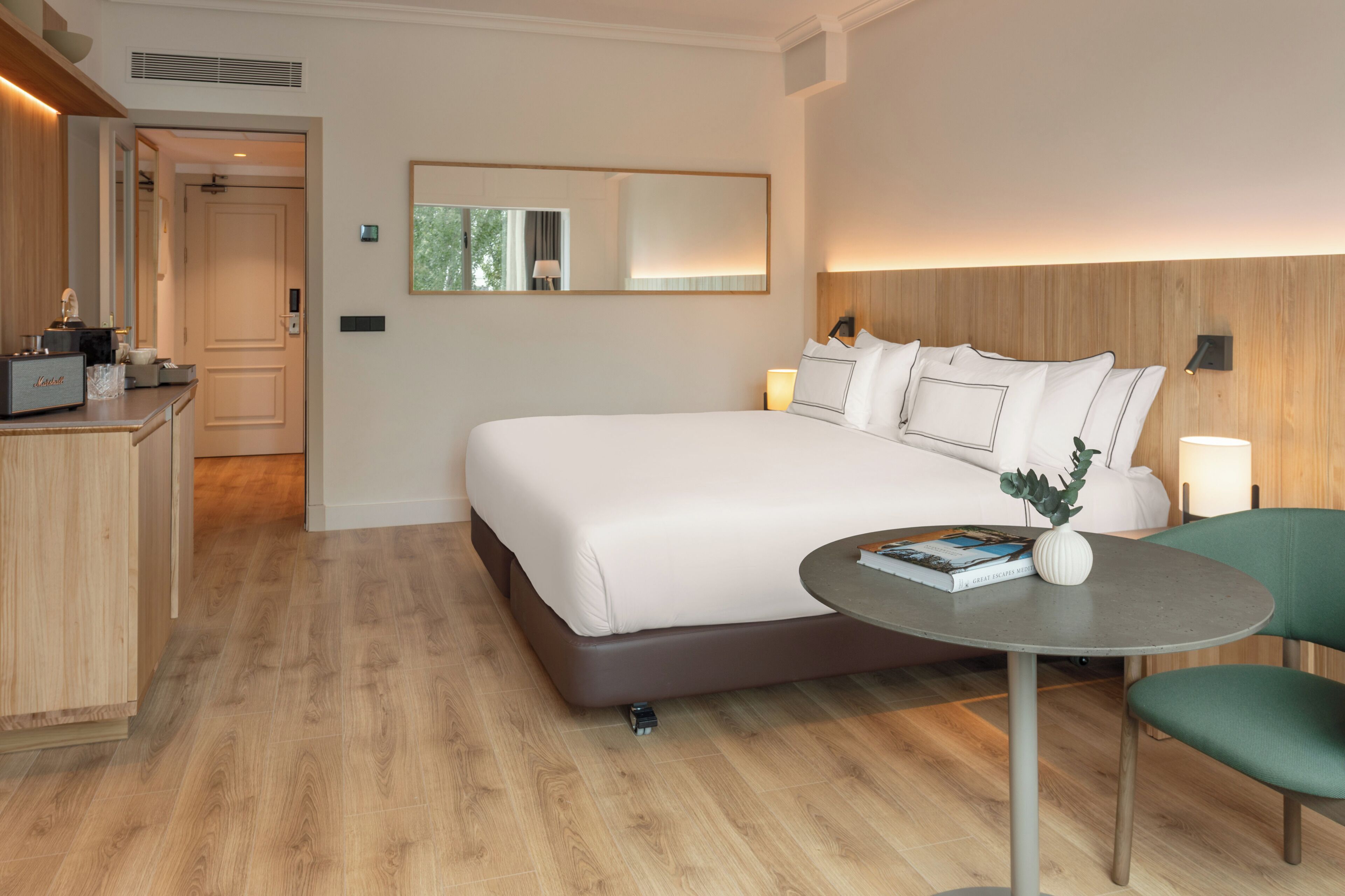 Ein modernes Hotelzimmer mit einem großen Bett, einem Nachttisch mit Lampe, einem kleinen runden Tisch mit Buch und Vase sowie einem grünen Sessel.