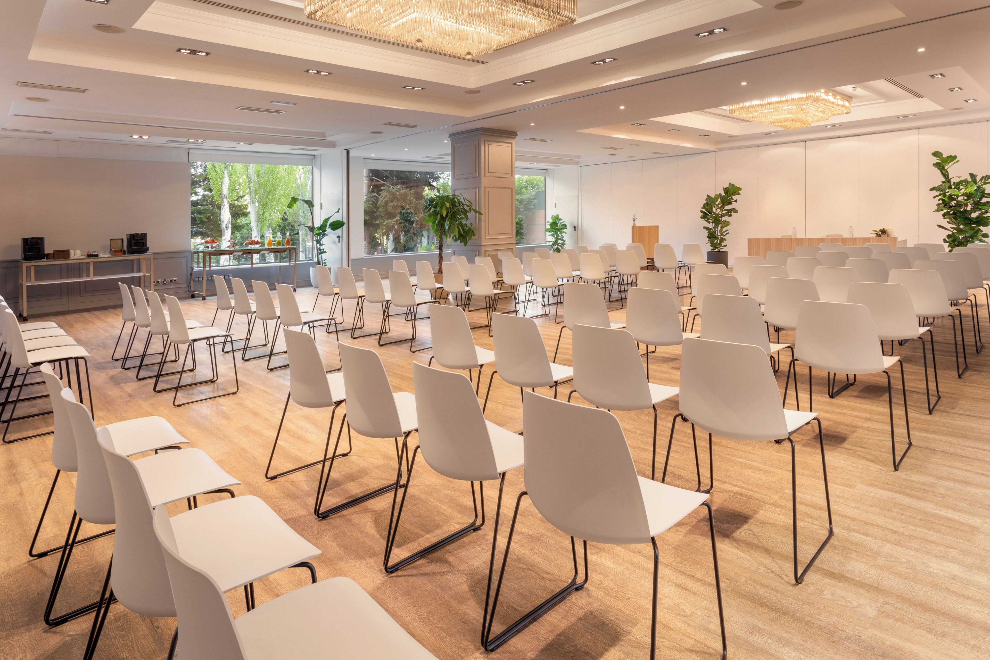 A conference room with rows of white chairs and tables, large windows with a view of trees, and a chandelier.