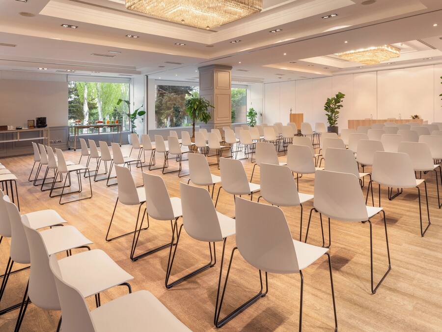 A conference room with rows of white chairs and tables, large windows with a view of trees, and a chandelier.