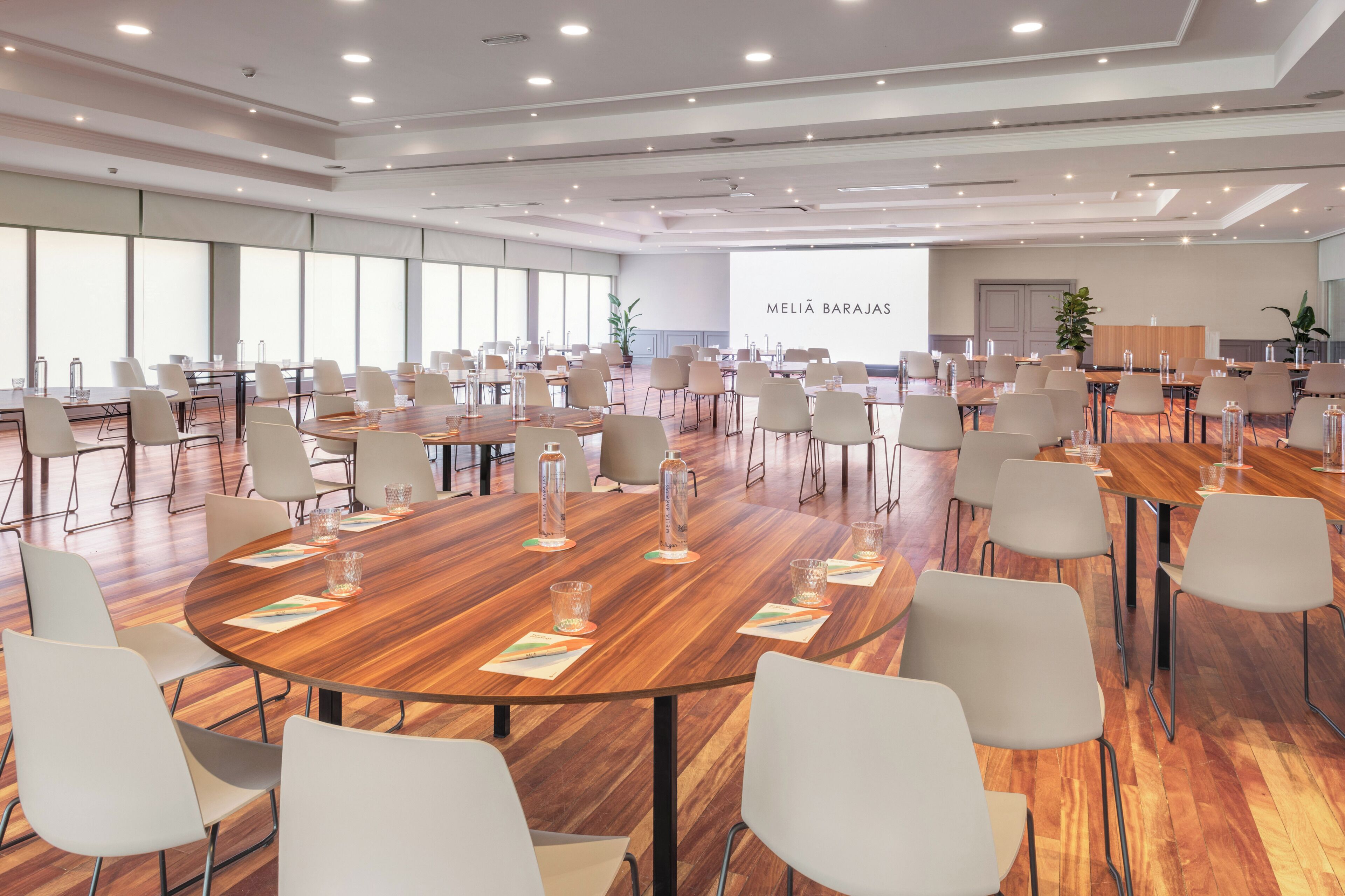Un salón de conferencias con varias mesas y sillas, ventanas amplias y un cartel con el letrero 'MELIA BARAJAS'.