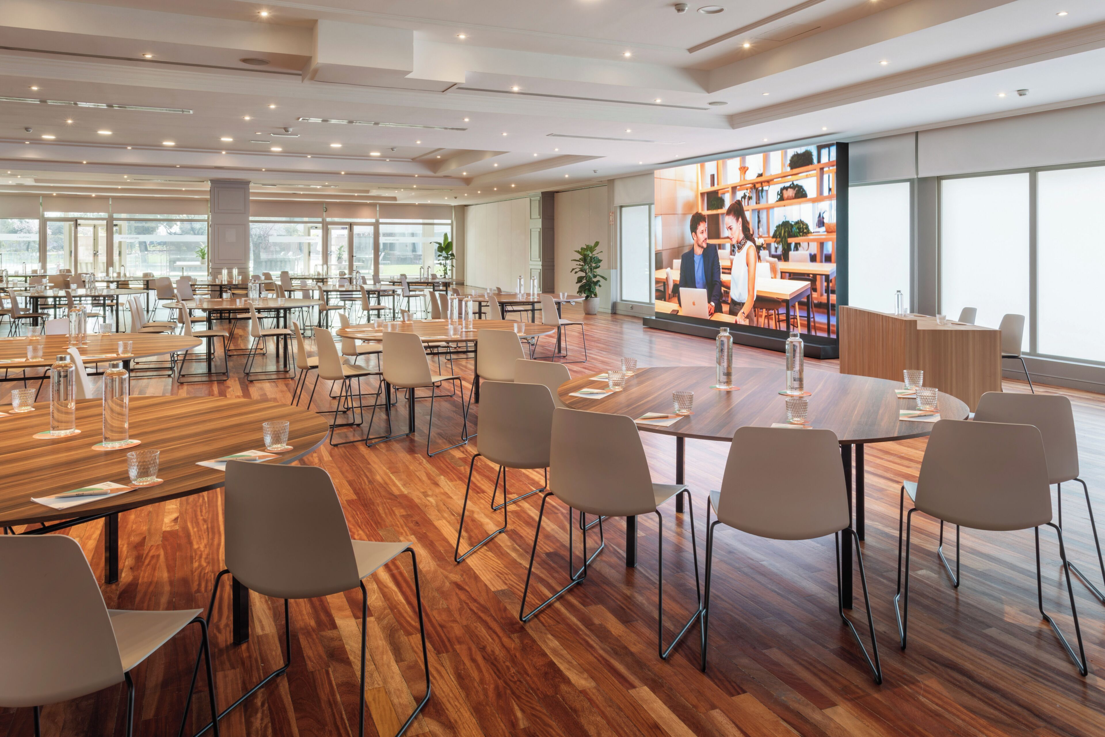 A spacious conference room with multiple tables and chairs, large windows, and a visible kitchen area.