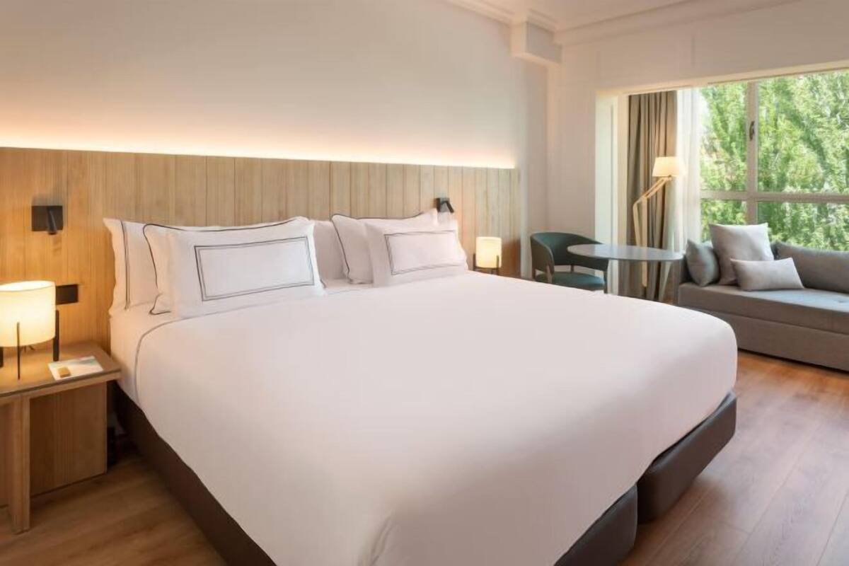 A hotel room with a large bed, a small table, a sofa, and a window with curtains.