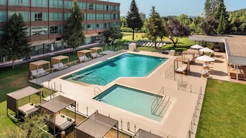 Una piscina rectangular con cubierta de madera, rodeada de sillones de descanso y sombrillas, con un edificio moderno y árboles al fondo.