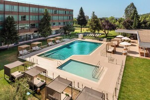 Ein rechteckiger Pool mit Holzterrasse, umgeben von Liegestühlen und Sonnenschirmen, vor einer modernen Gebäudeecke und Bäumen.