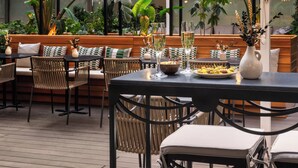 Un espacio al aire libre con bancos y sillas de madera, una mesa con un cuenco de fruta y un florero con flores.