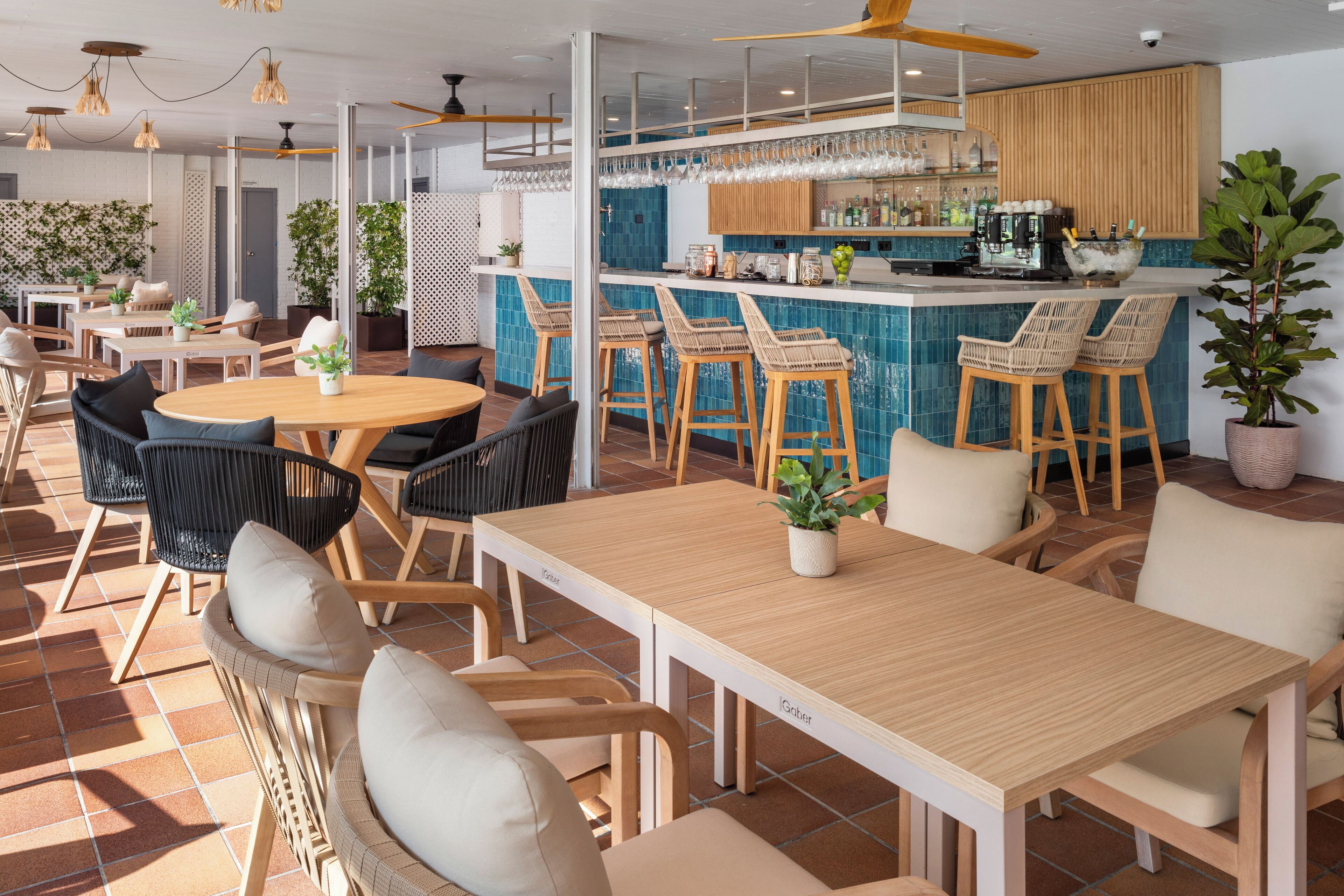 A modern restaurant with a bar area, wooden tables and chairs, and potted plants.