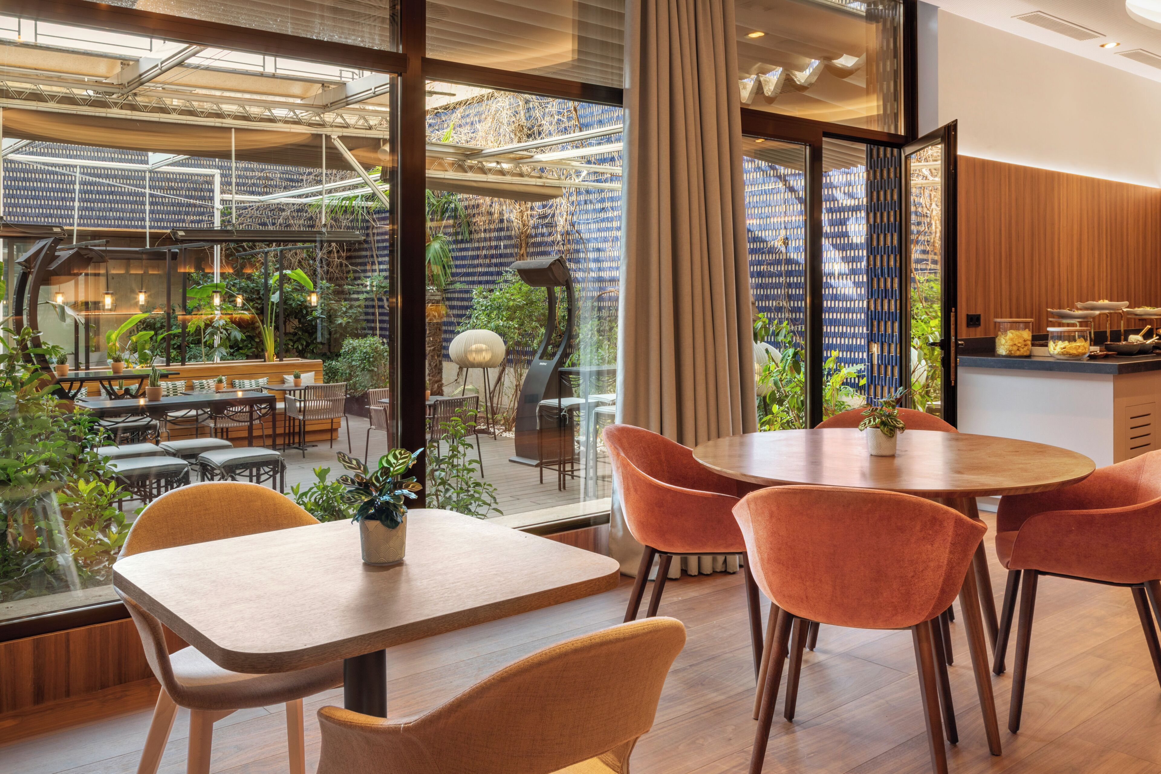 Un comedor moderno con mesas de madera y sillas naranjas, un separador de vidrio y vistas a un jardín exterior.