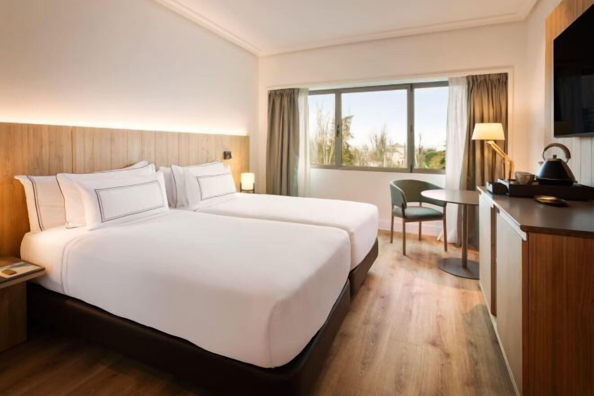 A hotel room with two beds, a desk, a chair, a TV, and a window with curtains.