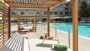 Un área junto a la piscina con sillones de descanso, una pérgola de madera y una mesa con una toalla y una bebida.
